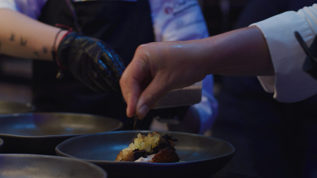 The photograph shows a close-up of the chefs' hands in action, where Alejandra Toloza's hand is seen meticulously assembling Anibal Almarza's "Jardín Umami" dish. The dish, which is part of the UmamiLab menu, consists of cabbage in herbs and Raise, adorned with crispy basmati rice and smooth cashew velvet, presented on a dark ceramic plate that enhances its colors and textures. The attention to detail suggests the precision and care put into the preparation of a culinary experience that highlights the umami flavor.