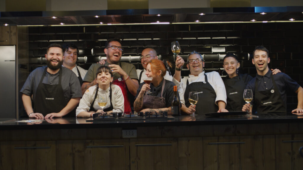The photograph shows the chefs of UmamiLab 2023, smiling and sharing a moment of camaraderie in the kitchen. Fernando Ramos, Roberto Soto, Javier Miranda, Anibal Almarza, Cristian Vieira, Alejandra Soulskin, Manuel Balmaceda, Alejandra Toloza, and Francisca Albornoz appear in a row behind the kitchen bar, some with glasses of wine in hand, celebrating the success of their culinary event. The image conveys a sense of joy and collective satisfaction, reflecting the spirit of collaboration and passion for gastronomy that characterizes UmamiLab.